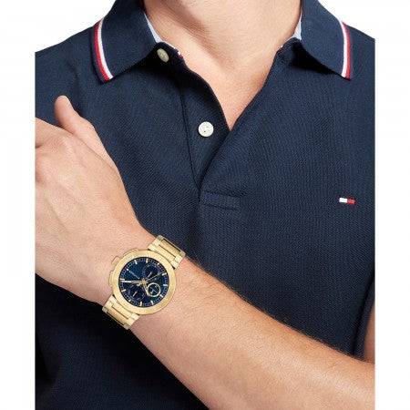 Tommy Hilfiger TH1792118 LORENZO Herenhorloge 44mm met goudkleurige band en blauwe wijzerplaat, gedragen bij navy poloshirt. Stijlvol accessoire voor herenmode.